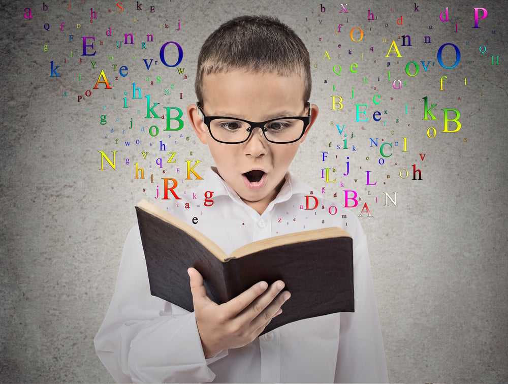 Surprised child reading a book with letters flying away from it isolated on grey wall background. Face expression. Education concept-1