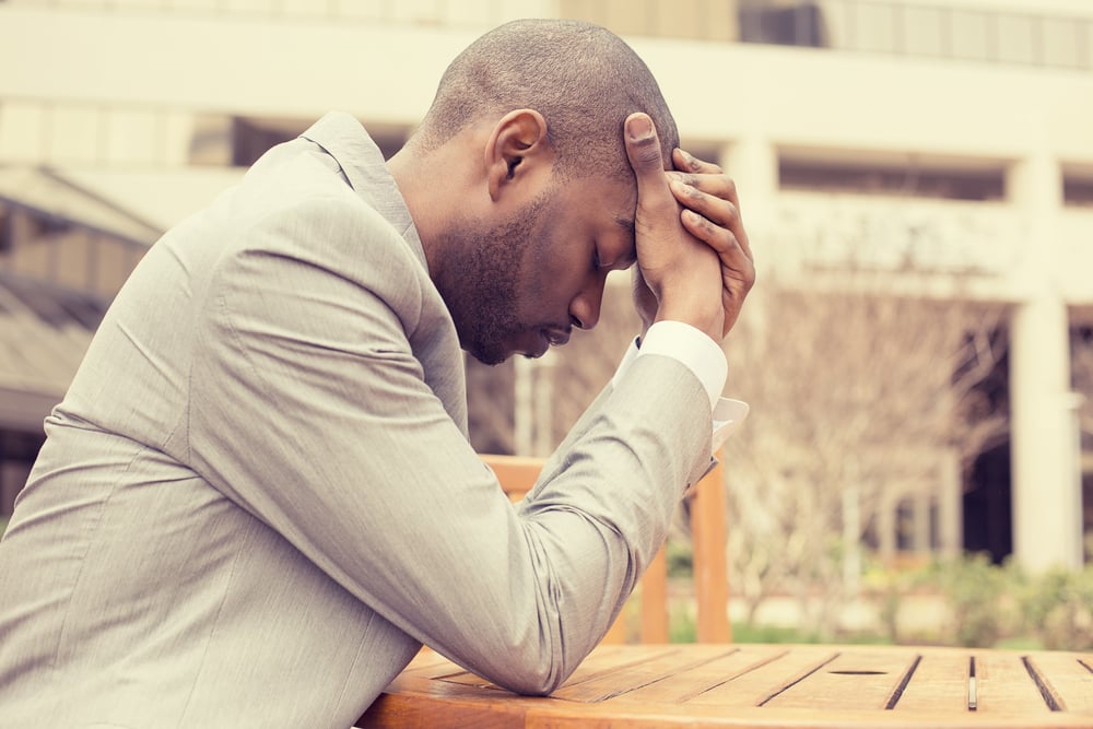 side profile stressed young businessman sitting outside corporate office holding head with hands looking down. Negative human emotion facial expression feelings.